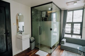 Un baño moderno con cabina de ducha de vidrio, un lavamanos blanco y un sofá gris en la zona de estar contigua.