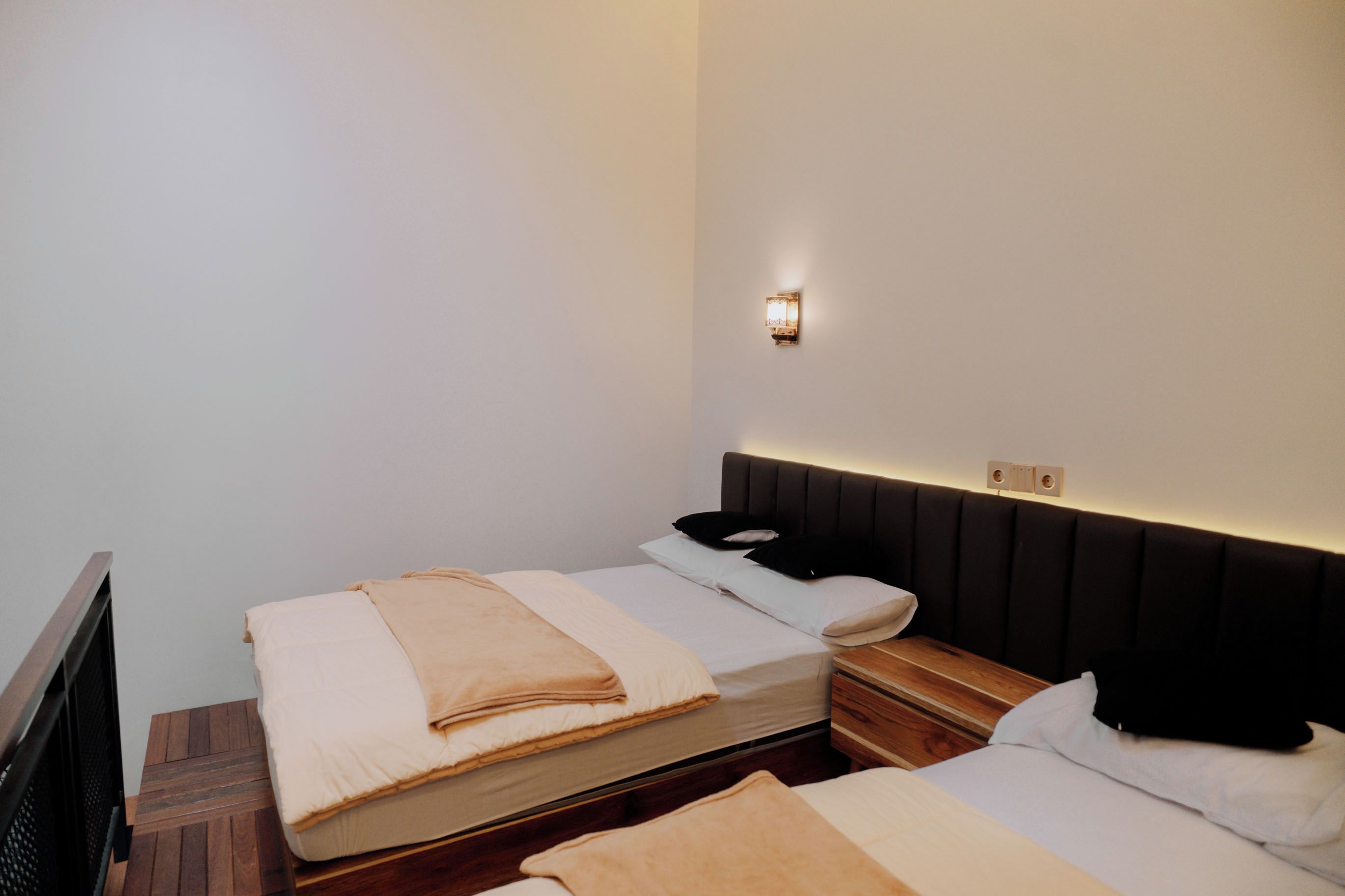 Quarto de hotel com duas camas, uma mesa de cabeceira, uma luminária na parede e piso de madeira.