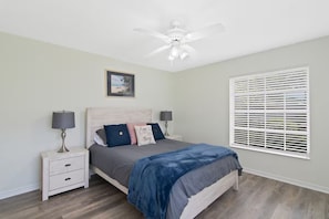 Quarto com cama, mesas de cabeceira, ventilador de teto, janela com persianas e um quadro na parede.