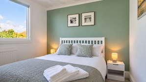 A neatly made bed with a white headboard, two bedside lamps, and a window with a view of greenery.