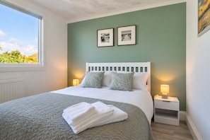 A neatly made bed with a white headboard, two bedside lamps, and a window with a view of greenery.
