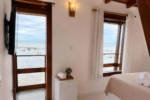 A bedroom with a bed, a small white cabinet, a wall-mounted TV, and a window with a view of the beach. - Pousada Villa Grauçá (Cairu)