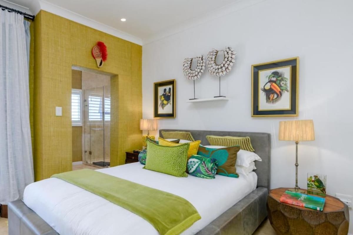 Habitación de hotel con una cama grande, una mesita de noche con lámpara, una pequeña mesa auxiliar con libros y un área de ducha.