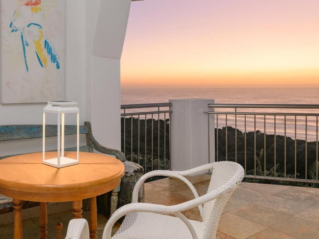 Un balcon avec vue sur la mer au coucher du soleil, une chaise en osier blanche, une table en bois avec une lanterne blanche et un tableau accroché au mur.