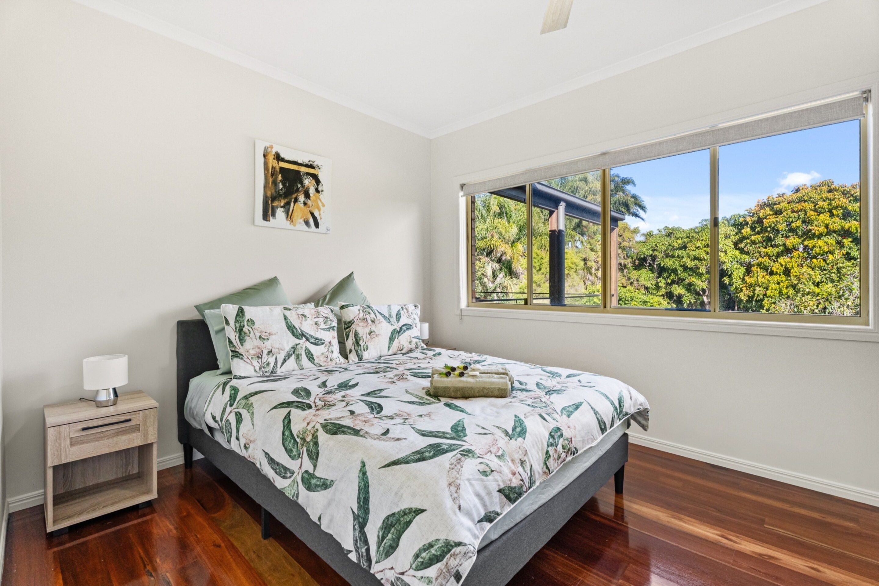 A bedroom with a bed, a nightstand, a lamp, a window with a view of trees, and a painting on the wall.