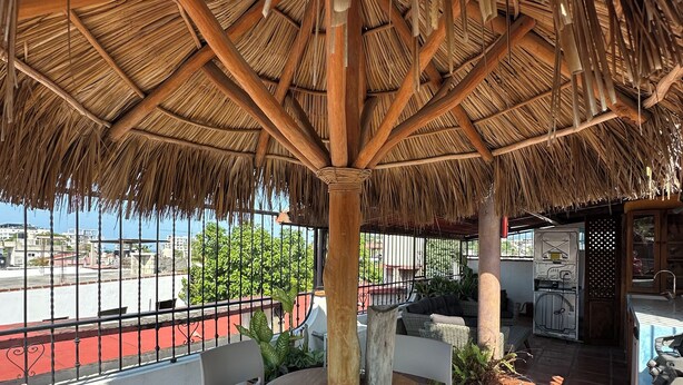 Een terras buiten met rieten dak, witte kunststof stoelen, een ronde tafel en uitzicht op de stad.