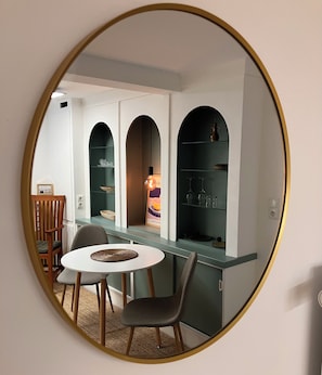 Una habitación con un espejo redondo que refleja un comedor, una cocina con estante empotrado y una pequeña mesa con sillas.