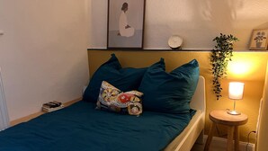 Uma cama com roupa de cama verde-água, uma almofada estampada, uma pequena mesa de cabeceira com abajur e um relógio de parede.