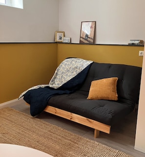 Una habitación con un sofá de color oscuro, una manta con estampado, un solo cojín marrón y una alfombra tejida.