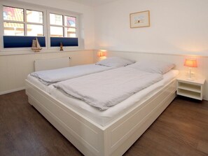Ein Doppelbett mit weißem Kopfteil und Fußteil, zwei Nachttische mit Lampen, ein Fenster mit Blick auf Gebäude und ein gerahmtes Bild an der Wand.