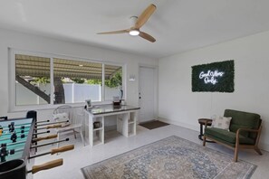 A room with a foosball table, a desk, a chair, a green sofa, a ceiling fan, and a neon sign. - Seacrest Bungalow - Cozy, Family, Beach @ 10 min (Largo)