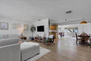 Uma sala de estar moderna com sofá, canto de jantar com mesa e cadeiras, e cozinha ao fundo.