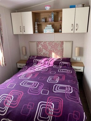 A bedroom with a bed, nightstands, a dresser, and a shelf with various items. - Cayton Bay Caravan Park - Holiday Accommodation 11337 (Scarborough)