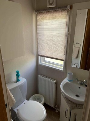 A compact bathroom with a toilet, a sink, a window with blinds, and a radiator. - Cayton Bay Caravan Park - Holiday Accommodation 11337 (Scarborough)