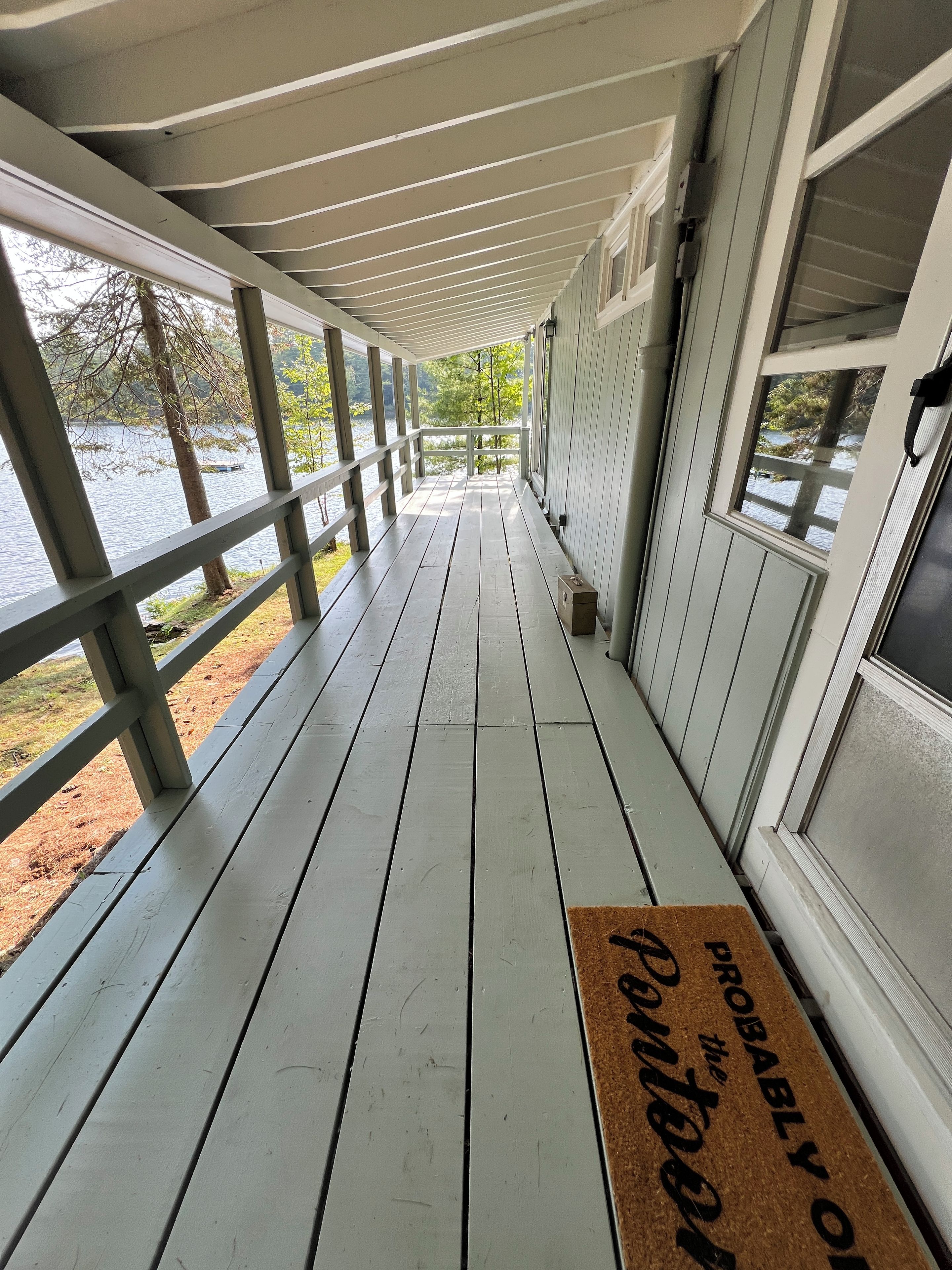 Une terrasse en bois couverte, avec vue sur un lac, un paillasson et une porte dotée d’une fenêtre.