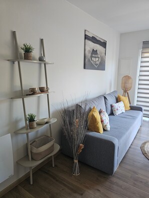 A living room with a grey sofa, wooden floor, and a wall-mounted shelf with decorative items. - Apartment 'Au Martin Pêcheur Côté Rivière' with Wi-Fi (Plobsheim)