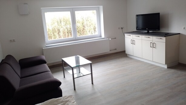 Sala de estar con un sofá, una mesa de centro de vidrio, un televisor sobre un mueble bajo y una ventana con vistas a árboles.