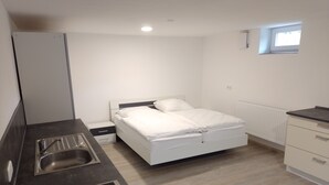 Un dormitorio compacto con una cama individual, una mesita de noche pequeña, un armario empotrado, una ventana y un área de kitchenette con lavabo y estufa.