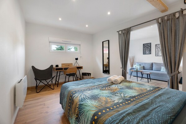 A modern bedroom with a large bed, a desk, and a chair. There is a window, a mirror, and a view of a balcony.