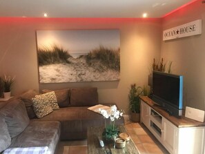 A living room with a large painting of a beach, a sofa, a coffee table with decorative items, a flat-screen TV, and a sign that reads 'OCEAN HOUSE'. - Apartment in Westerland With Swimming Pool (Sylt)