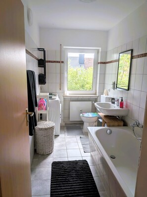 Ett badrum med badkar, toalett, handfat och ett fönster med utsikt över grönområden utanför.