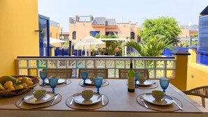 A dining table set with blue glassware and plates, a bottle of wine, and a bowl of fruit on a balcony with a view of buildings and greenery. - Vrbo Property (Sonneland (Urbanizacion))