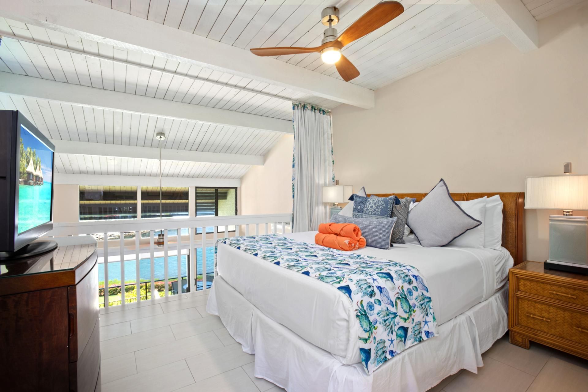 A hotel room with a bed, a TV, a ceiling fan, and a view of the ocean.