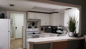 A kitchen with white cabinets, a stove, a microwave, and a coffee maker. There is a plant on the counter and a door with a 'HOME' sign. - Three Rivers Retreat (Lake City)