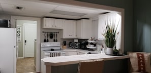 A kitchen with white cabinets, a stove, a microwave, and a coffee maker. There is a plant on the counter and a door with a 'HOME' sign. - Three Rivers Retreat (Lake City)