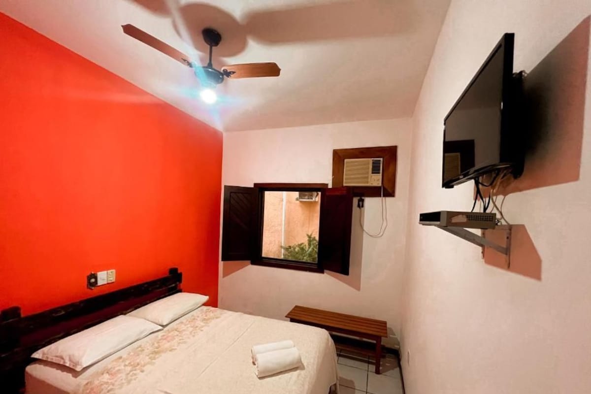 A bedroom with a bed, a ceiling fan, a TV mounted on the wall, and a window with an air conditioning unit.