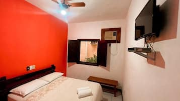 A bedroom with a bed, a ceiling fan, a TV mounted on the wall, and a window with an air conditioning unit.