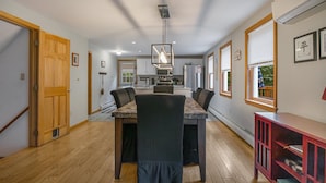 A dining area with a wooden table and chairs, a hanging light fixture, and a kitchen in the background.