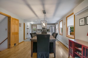 A dining area with a wooden table and chairs, a hanging light fixture, and a kitchen in the background.