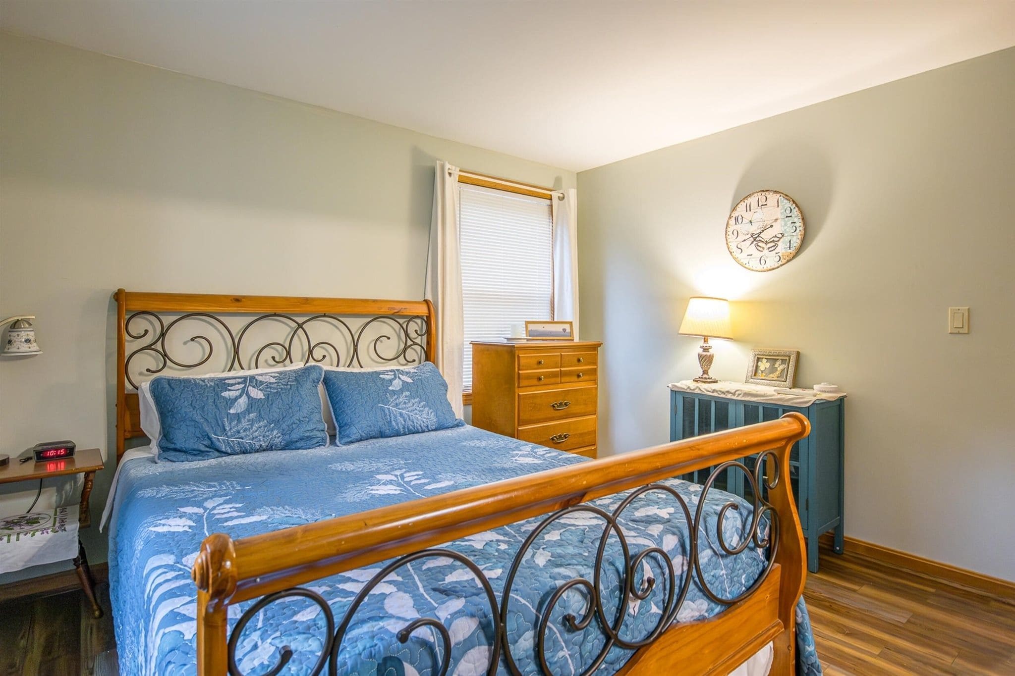 A bedroom with a wooden bed, a dresser, a bedside table with a lamp, and a wall clock.