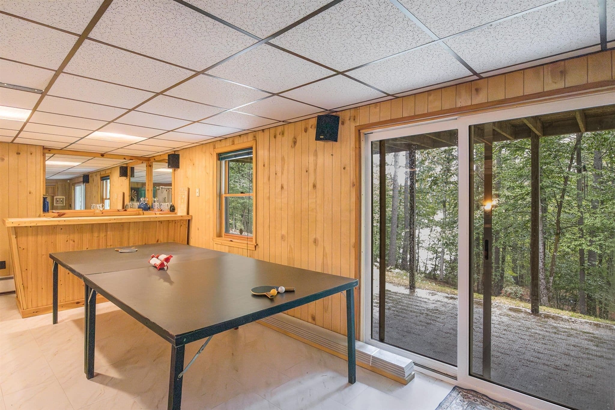 A room with a ping pong table, a bar area, and a large sliding glass door leading to a forested area.