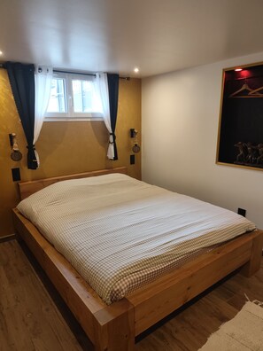 Une chambre à coucher avec un lit en bois, une fenêtre avec des rideaux, un luminaire fixé au mur et un tableau encadré.