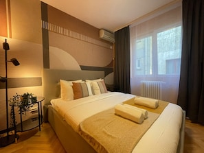 A hotel room with a large bed, a bedside table with a lamp, a potted plant, and a view of the outside through a window. - Chill Flow Calea Victoriei next to The Old Town (București)