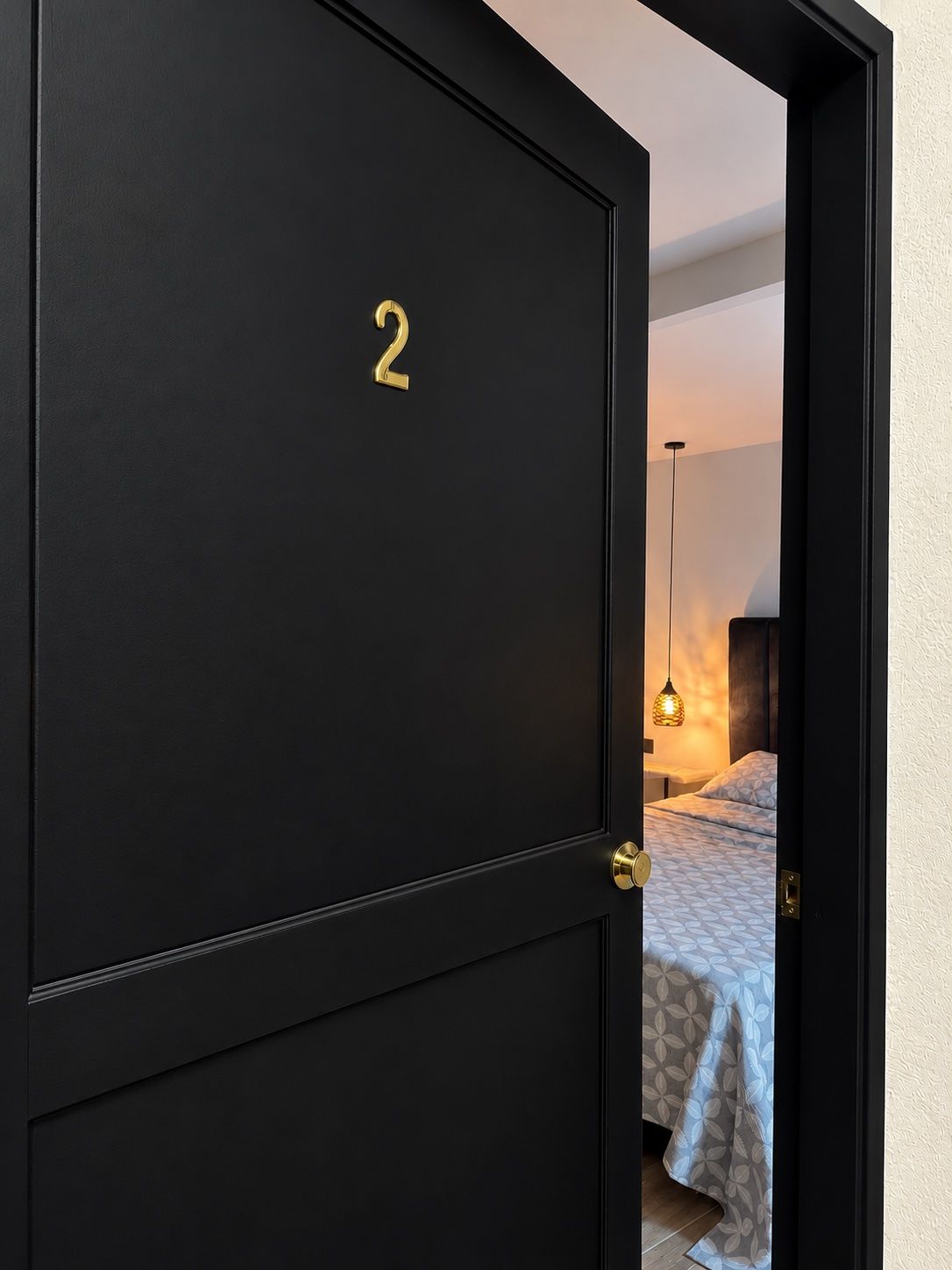 A black door with the number 2, leading to a bedroom with a bed and a hanging light fixture.