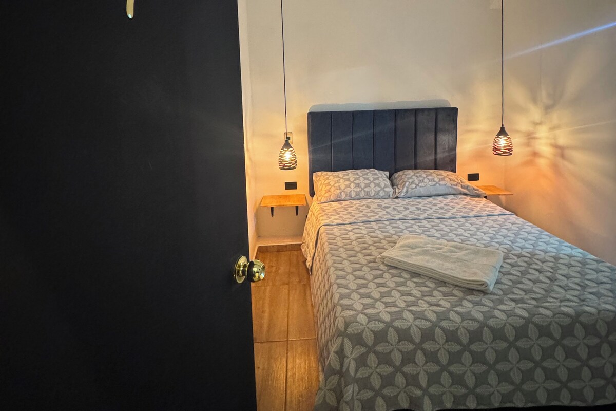 A bedroom with a bed, two bedside tables, a nightstand, and two pendant lights.