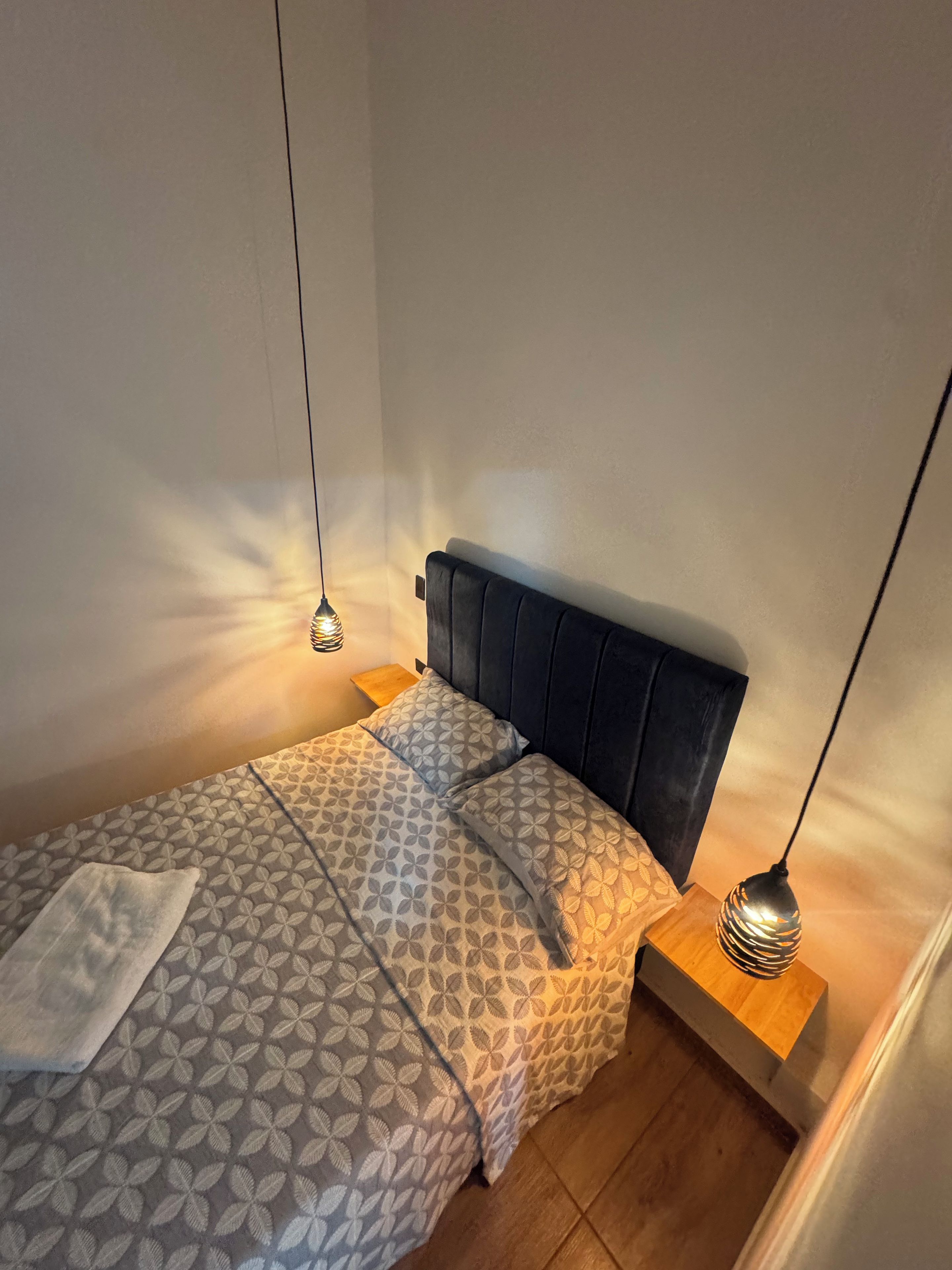 Un lit simple avec une couvre-lit à motifs et des oreillers, une tête de lit sombre et deux lampes de chevet posées sur des tables de chevet en bois.