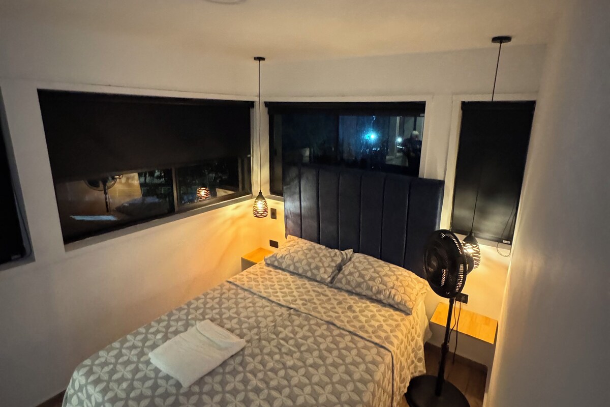 A bedroom with a bed, bedside lamps, a standing fan, and a view through the window.