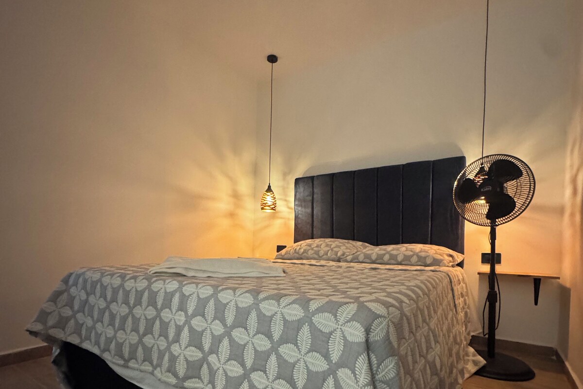 A bedroom with a bed, a bedside table, a wall-mounted fan, and a hanging light fixture.