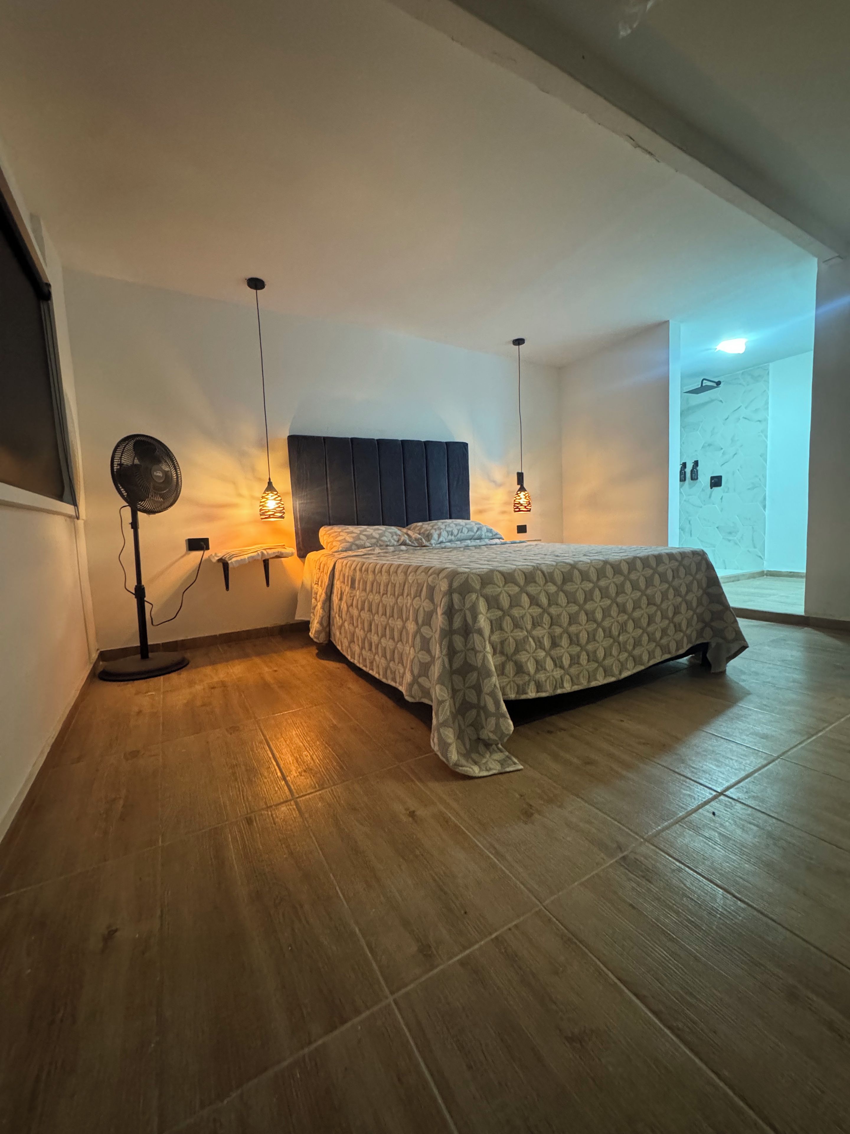 Une chambre à coucher avec un lit, des lampes de chevet, un ventilateur sur pied et une vue sur une porte.