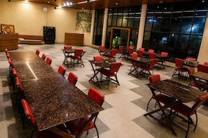 A spacious indoor dining area with multiple tables and chairs, a reception desk, and large windows. - Royal Reef Resort (Depalpur)