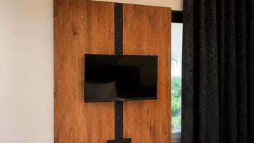 A modern living room with a flat-screen TV mounted on a wooden panel wall, a black shelf below, and a window with black curtains.