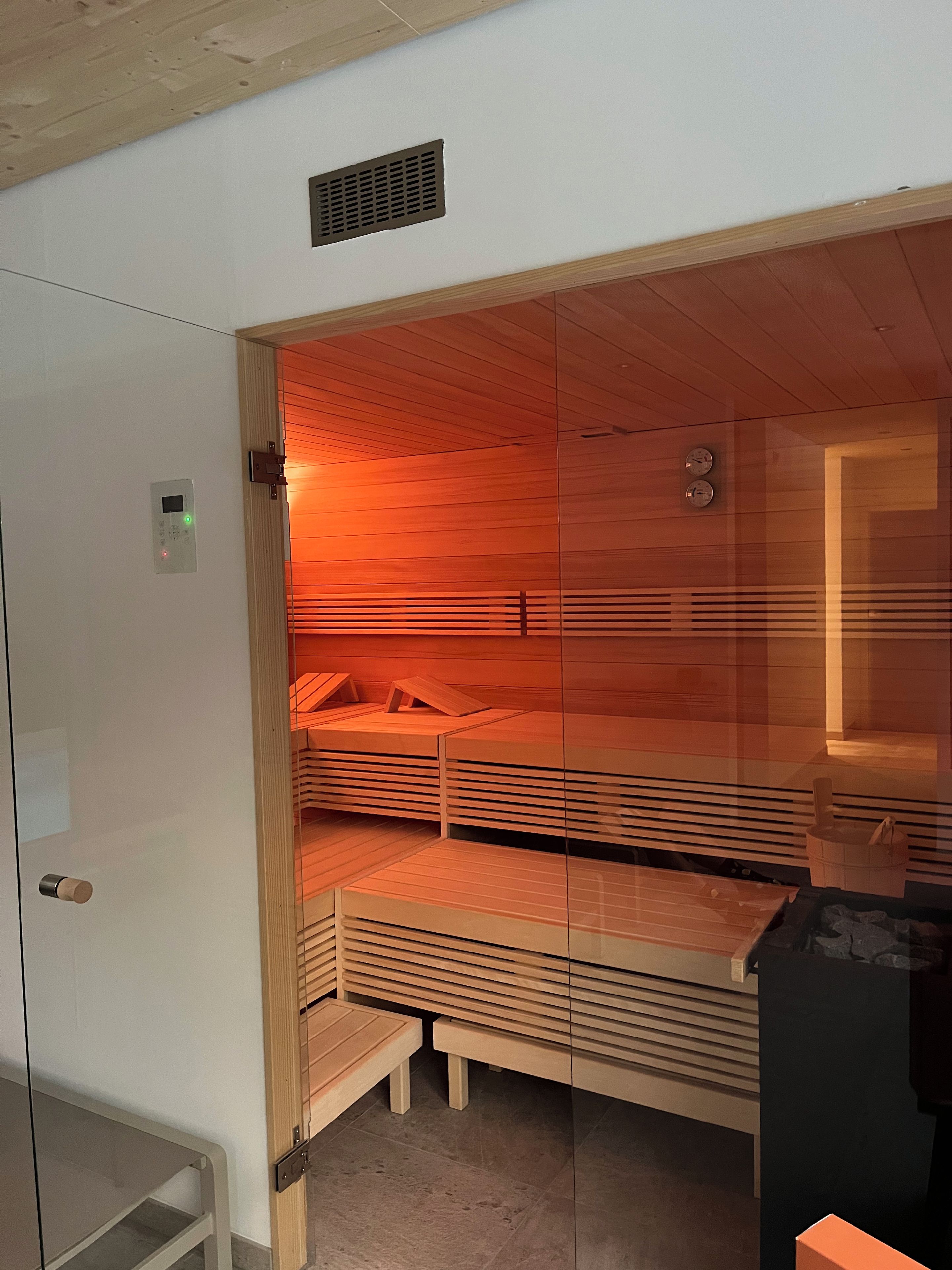 A sauna with wooden benches and a glass partition, a control panel, and a clock on the wall.