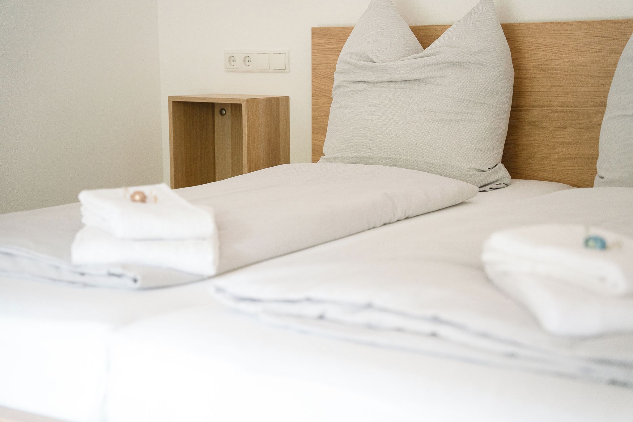 Una cama bien hecha con sábanas y almohadas blancas, un cabecero de madera y una mesita de noche de madera con una lámpara.