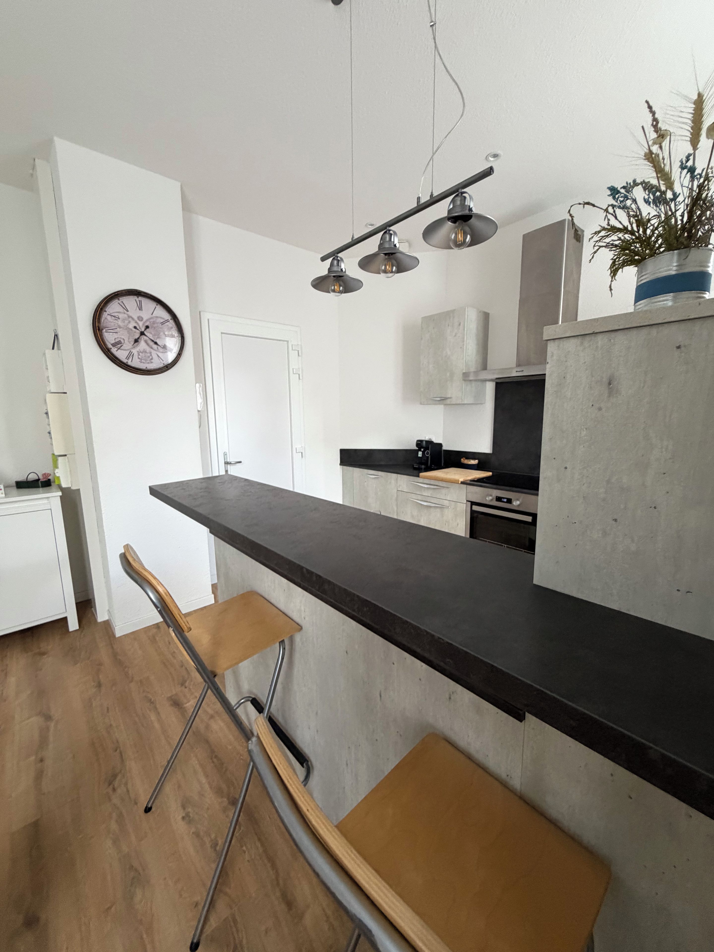 A modern kitchen with a long countertop, two bar stools, a clock, and pendant lighting.