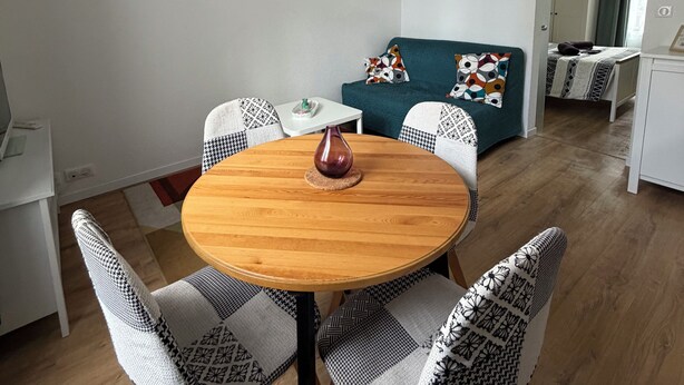 A modern living room with a round wooden table, patterned chairs, a teal sofa, and a small white side table. - Charmant T2 avec Terrasse et Jardin - Le Gamay (Châtel-Guyon)