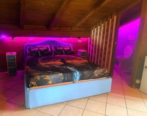 Una camera da letto con un letto, travi in legno a soffitto, pavimento piastrellato e una parete illuminata di viola.